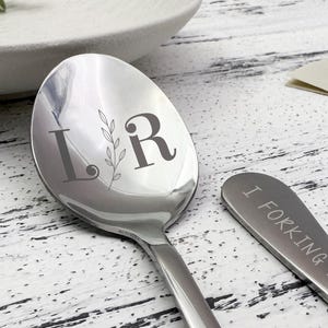 May include: A stainless steel spoon with the initials "L R" and a sprig of leaves engraved on the bowl. A smaller spoon with the phrase "I FORKING LOVE YOU" is also visible.