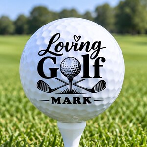May include: A white golf ball on a white tee featuring black printed text that reads Loving Golf with a small heart icon above the word Loving. Below the text is a graphic of a golf ball on a tee with two crossed golf clubs and the name MARK.