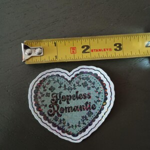 May include: A heart-shaped sticker with the words "Hopeless Romantic" in a decorative font. The sticker has a light blue background with floral accents and a glittery border. The sticker is approximately 2 inches wide.