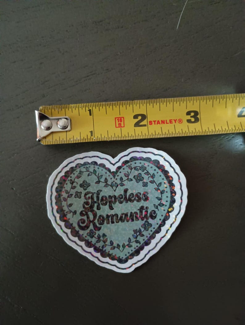 May include: A heart-shaped sticker with the words "Hopeless Romantic" in a decorative font. The sticker has a light blue background with floral accents and a glittery border. The sticker is approximately 2 inches wide.