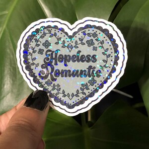 May include: A heart-shaped sticker with the words "Hopeless Romantic" in a script font. The sticker has a floral border and a holographic, glittery effect. The background is a green leafy plant.