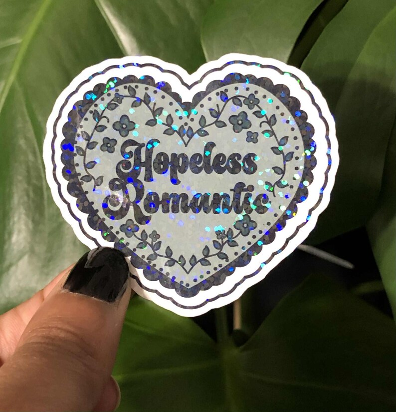 May include: A heart-shaped sticker with the words "Hopeless Romantic" in a script font. The sticker has a floral border and a holographic, glittery effect. The background is a green leafy plant.