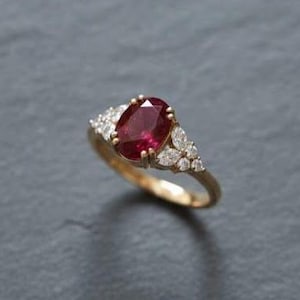 以下が含まれることがあります： 大きな楕円形のルビーを中央に配したゴールドリング。ルビーは深い赤色をしています。ルビーの両側には、小さな透明な宝石がセットされ、輝きを添えています。リングはゴールドのバンドにセットされています。