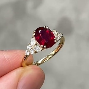 以下が含まれることがあります： 大きな楕円形のルビーを中央に配したゴールドリング。ルビーは深い赤色で、より小さな透明な宝石に囲まれています。リングのバンドは磨かれたゴールドで、セッティングは繊細で装飾的です。