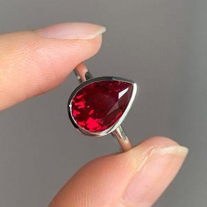 Könnte beinhalten: Ein silberner Ring mit einem tropfenförmigen, tiefroten Edelstein. Der Edelstein ist in einer Lünettenfassung gefasst. Der Ring wird zwischen zwei Fingern vor einem grauen Hintergrund gehalten. Der Ring hat ein dünnes, poliertes Band.