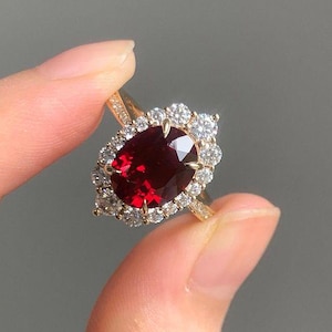 May include: A gold ring featuring a large, oval-cut, deep red gemstone surrounded by a halo of smaller, clear gemstones. The ring is held up, showcasing its intricate design and sparkle. The setting appears to be gold.