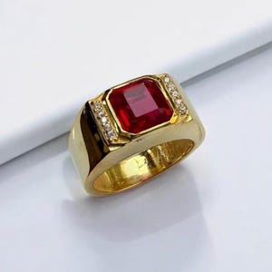 May include: A gold-colored ring with a large, rectangular, red gemstone at the center. The ring is accented with small, clear stones on each side of the central stone. The ring is set on a white surface.