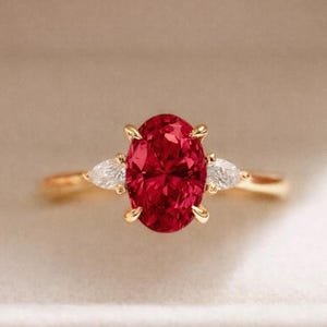 May include: A gold ring featuring a large, oval-cut ruby gemstone as the centerpiece, flanked by two smaller pear-shaped diamonds. The ring is set in a classic three-stone design, displayed in a white jewelry box.