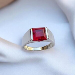 Puede incluir: Un anillo de plata con una llamativa gema roja brillante de corte cuadrado. El anillo tiene un diseño minimalista con un acabado brillante. La gema es la característica principal, engastada en un bisel tradicional. El anillo se muestra sobre un fondo blanco.