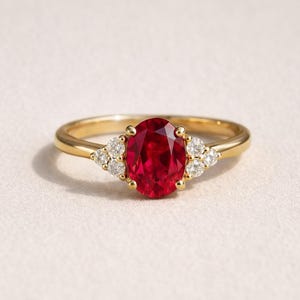 May include: A gold ring featuring a large, oval-cut ruby gemstone as the centerpiece. Flanking the ruby are clusters of smaller, round, white gemstones. The ring has a delicate band and a classic, elegant design.