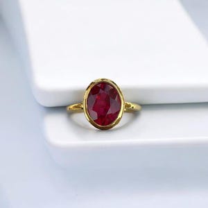 May include: A gold ring with a large, oval-cut ruby gemstone. The ruby is a deep red colour and is set in a gold bezel. The ring has a simple, elegant design.
