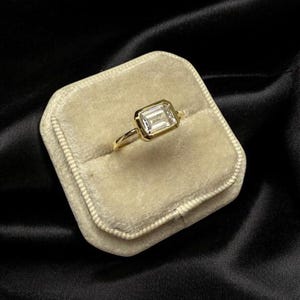 May include: A gold ring featuring a rectangular, clear gemstone in a bezel setting. The ring is presented in a beige velvet ring box against a black backdrop.