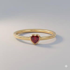 May include: A gold ring with a heart-shaped, red gemstone. The ring has a thin band and a bezel setting for the gemstone. The heart-shaped stone is a vibrant red color.