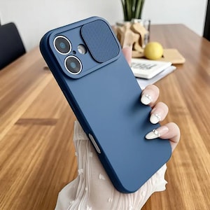 Puede incluir: Una funda de teléfono azul marino con una cubierta para la lente de la cámara. La funda tiene un acabado mate y presenta dos recortes para la cámara y una cubierta deslizante. La funda del teléfono está sujeta por una mano con uñas cuidadas.
