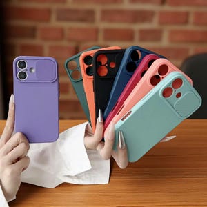 May include: A collection of colorful phone cases in various shades, including purple, green, orange, black, blue, pink, and teal. Each case features a camera lens cover. The cases are displayed against a brick wall and wooden surface.