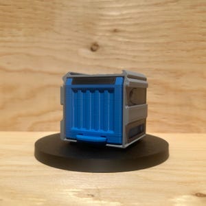 Peut inclure: Maquette de conteneur imprimée en 3D, bleue et grise, avec un dessus noir et une petite poignée. Le conteneur est posé sur une base noire circulaire, sur un fond en bois clair.