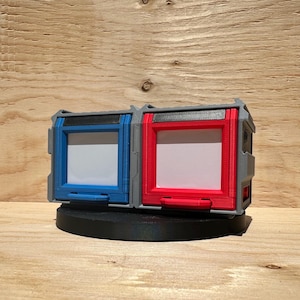 Peut inclure: Deux conteneurs imprimés en 3D, l'un bleu et l'autre rouge, avec des ouvertures rectangulaires blanches. Les conteneurs sont montés sur une base noire circulaire. Le design comprend des accents gris et un style futuriste.