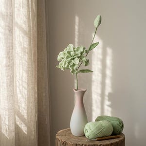 以下が含まれることがあります： 長い茎と閉じた蕾を持つライトグリーンのクロッシェ編みの花が、滑らかなグラデーションカラーの花瓶に飾られています。花瓶の横には、ライトグリーンの毛糸玉2つと木製のクロッシェ針が、質感のある木製の表面の上に置かれています。