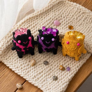 Op de afbeelding: Drie kleine, gehaakte pluche figuren met ronde lichamen en lachende gezichten. Twee zijn zwart met roze of paarse accenten en kristalachtige stenen bovenop, de derde is geel met barnsteenkleurige stenen bovenop.