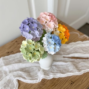 May include: A bouquet of crocheted hydrangea flowers in various colors including light purple, soft pink, pale green, white, light blue, yellow, and orange. Each flower head is composed of multiple small, textured petals with yellow centers.