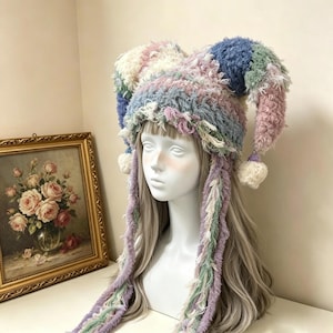 May include: A multi-colored jester-style hat crafted from fuzzy, textured yarn in shades of blue, pink, green, and cream. The hat features two long, pointed ear-like sections with cream-colored pom-poms at the ends and long, dangling fringe.