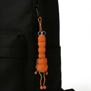 May include: A handmade orange crochet charm, resembling a quirky creature with black eyes and dangling legs, hangs from a black backpack zip. The charm's design is playful and unique, adding a touch of whimsy to the backpack.