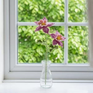 以下が含まれることがあります： 黄色い中心部を持つ紫とピンクの2つの花、緑の葉、細い緑の茎を備えたかぎ針編みの花の茎。花は透明なガラスの花瓶に飾られており、複雑な糸の質感と詳細な花びらの形がはっきりと見て取れます。