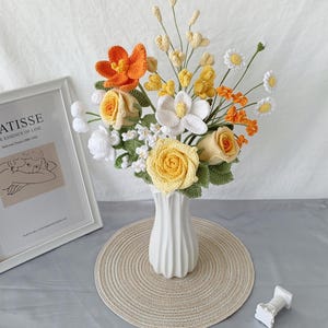 May include: A handcrafted crochet bouquet with yellow roses, orange lilies, white daisies, and other flowers in a white vase. The arrangement is on a beige placemat, with a framed print in the background.