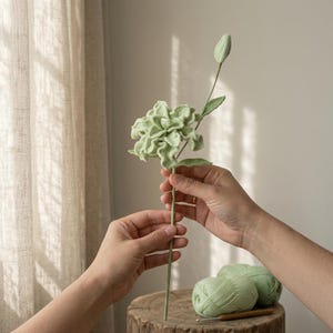 以下が含まれることがあります： 淡いセージグリーンのクロッシェ編みの花。長く細い茎に、閉じたつぼみ1つと小さな葉が2枚ついています。毛糸で編まれた複雑で重なり合う花びらが特徴で、細部まで作り込まれた質感のあるハンドメイドの仕上がりです。