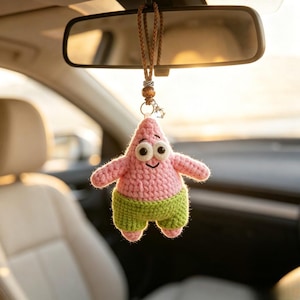 May include: A crocheted hanging ornament shaped like a pink starfish character with large white eyes and a smiling mouth. The figure wears green shorts and is suspended by a braided brown cord featuring a small wooden bead and a silver charm.