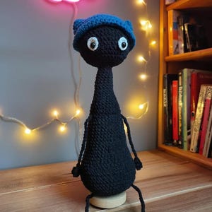 Può includere: Bambola nera all'uncinetto con berretto blu. La bambola ha grandi occhi bianchi e piccole braccia e gambe. È seduta su una base di legno. Lo sfondo include un fiore al neon rosa e una libreria.