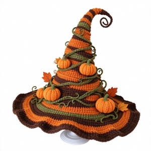 May include: A crocheted witch hat featuring horizontal stripes in orange, brown, and olive green. The pointed top curls at the end. The hat is decorated with small crocheted orange pumpkins, green vines, and fabric autumn leaves.