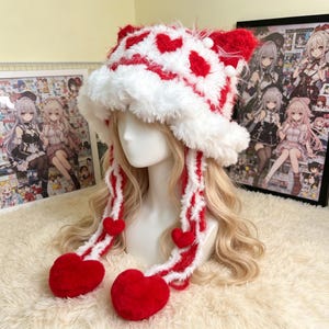 May include: A fuzzy white and red bucket hat featuring a repeating heart pattern, cat-like ear shapes on the crown, and two long braided side tassels ending in large, plush red heart-shaped pom-poms, displayed on a mannequin head.