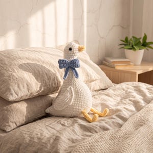 May include: A white crocheted goose plush toy featuring a yellow beak and feet, with a light blue crocheted bow tied around its neck. The goose has black button eyes and is crafted with a visible yarn stitch pattern throughout its body.