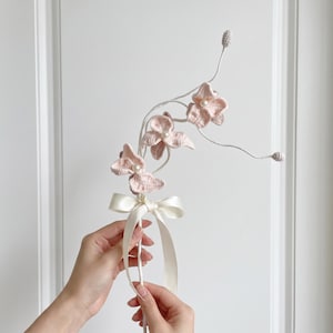以下が含まれることがあります： 淡いピンク色のクロッシェ編みの花が3つ付いた装飾用の茎。それぞれの花の中心には小さな白いパールがあしらわれています。茎は白い糸で巻かれ、先端には2つの小さなクロッシェ編みのつぼみがあり、クリーム色のサテンリボンの結び目で仕上げられています。