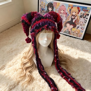 May include: A jester-style hat crafted from textured, fuzzy yarn in a variegated color palette of deep red, dark purple, and black. The hat features two long, pointed ear-like extensions with pom-poms at the ends and two long, dangling side panels.