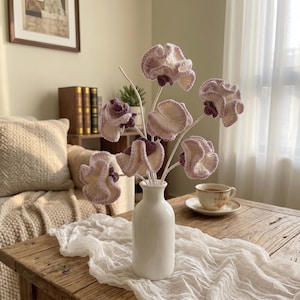 Pode incluir: Um buquê de seis flores de crochê com pétalas onduladas em tons de creme e roxo claro, presas a hastes brancas finas. As flores estão dispostas em um vaso de cerâmica branca lisa com um gargalo estreito e uma base arredondada.