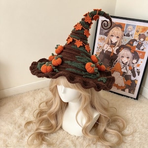 May include: A brown crocheted witch hat featuring a curled tip, decorated with small orange pumpkins and green crocheted leaves. The hat has a wide, wavy brim and is displayed on a mannequin head with long, wavy blonde hair.