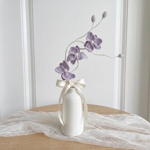 以下が含まれることがあります： 滑らかな質感の白い陶器製の花瓶に、クリーム色のサテンリボンが結ばれています。中には、中央に白いパールをあしらったラベンダー色のクロッシェ編みの花が3輪あり、細い白い茎の先に小さな質感のある蕾がついています。