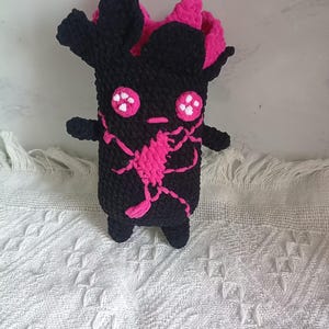 May include: A black crocheted plush toy with a cylindrical body, two small arms, and two short legs. The figure features bright pink accents, including a central spider-like graphic, two round eyes with white dots, a small mouth, and a pink interior top.