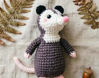 May include: A crocheted stuffed animal shaped like an opossum with a white face, dark gray body, black ears, and a light pink nose, feet, and tail. The creature has small black eyes and subtle pink blush marks on its cheeks, crafted with yarn.