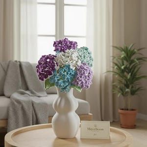 May include: A white vase displays a bouquet of crocheted hydrangeas in shades of purple, blue, and white. The flowers are on a round wooden table. A card with the text "Maya Bloom" is on the table. A sofa and a potted plant are in the background.