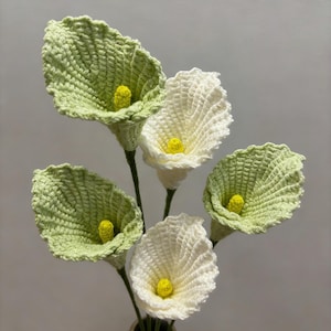 May include: A bouquet of handmade crochet calla lilies in a vase. The flowers feature green and white petals with yellow centers. The stems are green, and the vase is a neutral beige color.