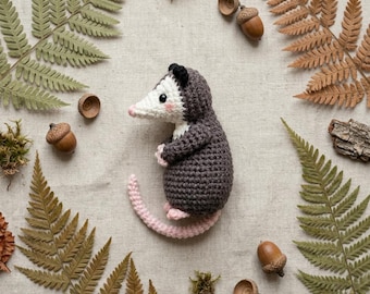 May include: A crocheted opossum toy with a dark gray body, a white face, black ears, and a light pink nose, feet, and tail. The opossum has small black eyes and pink blush details on its cheeks, crafted with a textured yarn stitch pattern.