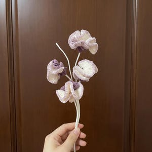 Pode incluir: Um caule de flor em crochê com quatro flores franzidas em tons de roxo claro e creme. As pétalas têm uma textura ondulada distinta com centros roxos escuros, presas a caules brancos, finos e flexíveis que se unem em uma base única.