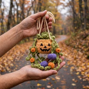 May include: A crocheted hanging ornament featuring a small orange jack-o'-lantern pot with a black stitched face, surrounded by green crocheted vines, small orange spheres, and two small purple spheres, suspended by a light beige yarn loop.