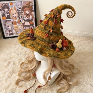 May include: A crocheted witch hat in variegated shades of green, yellow, and brown, featuring a curled tip, dangling crocheted leaves, and decorative clusters of small crocheted pumpkins, gourds, and pinecones attached to the wide, floppy brim.