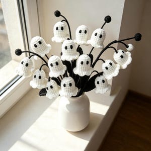 May include: A white ceramic vase containing a bouquet of crocheted ghost figures. Each ghost is white with two black embroidered eyes, attached to thin black stems with small black spheres at the tips and dark green crocheted leaves.