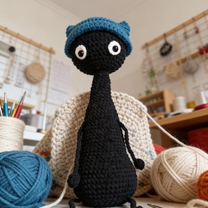 Könnte beinhalten: Handgefertigte schwarze Häkelfigur mit blauem Hut und großen weißen Augen. Die Figur hat einen langen, schlanken Körper und kleine Arme und Beine. Garn und Häkelzubehör deuten auf ein Handarbeitsprojekt hin.