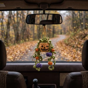 May include: A crocheted car hanging ornament featuring a light orange jack-o'-lantern pot with a black stitched face. Green crocheted vines trail downwards, accented with small orange and purple crocheted spheres hanging from the leafy strands.
